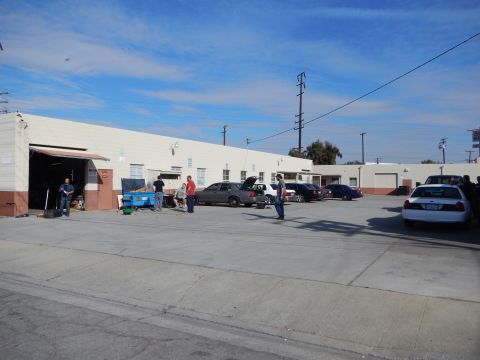2003 Ford Crown Victoria - People stand and walk near an open garage door of a long industrial warehouse, with sedans including a white one parked across the lot.