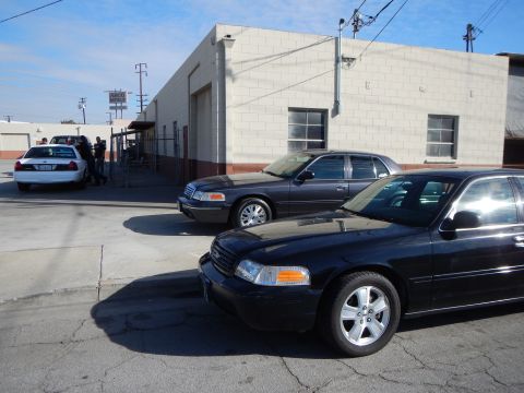 2003 Ford Crown Victoria - black Ford sedan in the foreground and a gray Ford sedan beyond it are parked on cracked pavement near a cinderblock building.