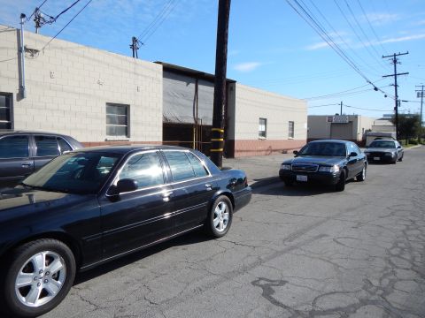 2003 Ford Crown Victoria - dark sedan in the foreground and more dark sedans parked down the block along an industrial street under a clear sky.