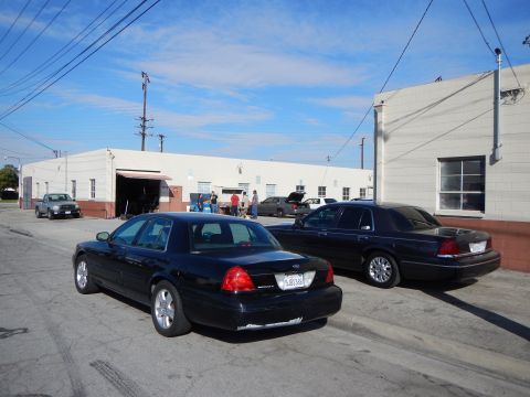2003 Ford Crown Victoria - Two dark Ford sedans, one showing the Ford rear emblem, are parked in a lot with people gathered by a warehouse door behind them.