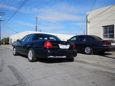 2003 Ford Crown Victoria - Two dark Ford sedans seen from behind, one with a visible California plate and Ford emblem, parked on pavement between buildings.