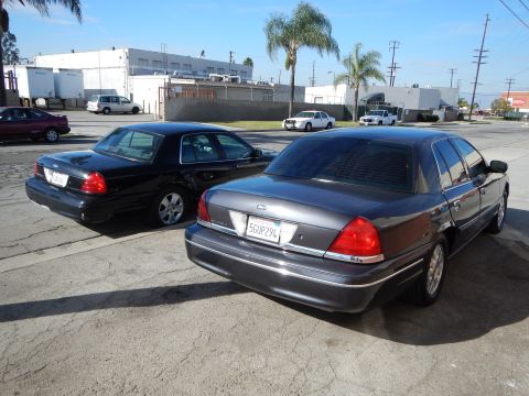 2003 Ford Crown Victoria - black and a gray Ford Crown Victoria sedan parked rear-first in an industrial lot with palm trees and warehouses behind them.