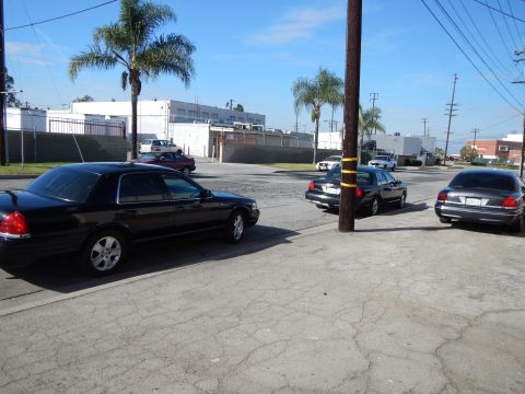 2003 Ford Crown Victoria - Several dark Ford sedans are parked at angles around a cracked-pavement industrial lot lined with palm trees and warehouses.