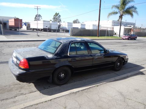 2003 Ford Crown Victoria - black Ford sedan with plain black steel wheels seen from the rear three-quarter, parked at a curb near fenced industrial yards.