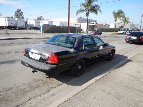 2003 Ford Crown Victoria - black Ford sedan on black steel wheels sits at a curb on cracked pavement, with palm trees and warehouses in the background.