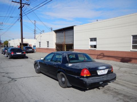 2003 Ford Crown Victoria - weathered black Ford sedan with steel wheels parked at a curb along an industrial street, with more dark sedans down the block.