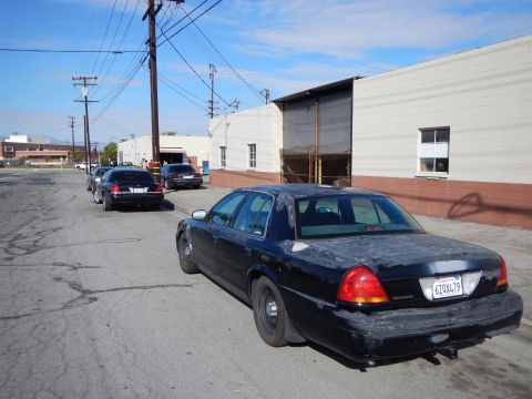 2003 Ford Crown Victoria - dark blue Ford sedan with steel wheels and a California plate parked at a curb on an industrial street with warehouses behind.