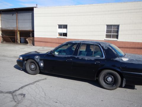 2003 Ford Crown Victoria - Side profile of a dark Ford sedan on steel wheels parked against a cinderblock industrial building under a clear blue sky.