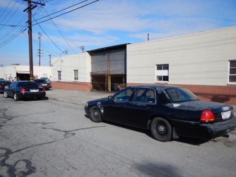 2003 Ford Crown Victoria - dark blue full-size sedan sits on a cracked asphalt lot beside beige industrial warehouse buildings under a clear sky.