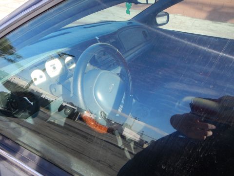 2003 Ford Crown Victoria - Looking through a windshield at a sedan's gray dashboard, steering wheel and instrument gauges with a hand reflected in the glass.