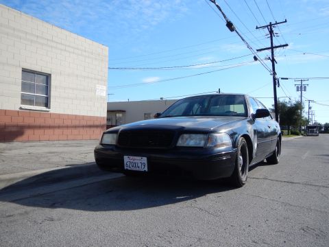 2003 Ford Crown Victoria - Low-angle front view of a black Ford sedan with California plates parked on asphalt beside a beige industrial building.