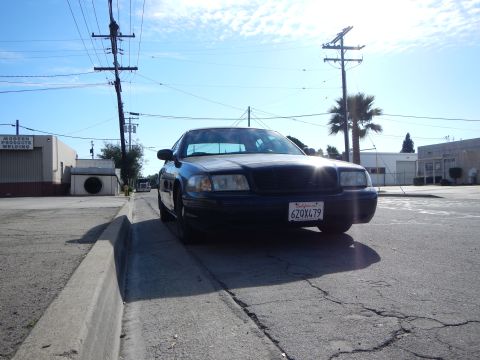 2003 Ford Crown Victoria - Head-on view of a black Ford sedan parked along a curb with a palm tree, power poles and warehouses in the background.