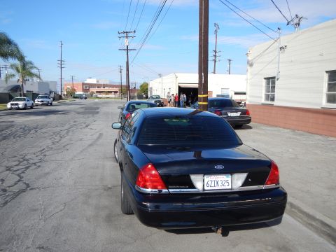2003 Ford Crown Victoria - Rear view of a dark Ford sedan with a California plate parked on an industrial street with power poles and warehouses behind.