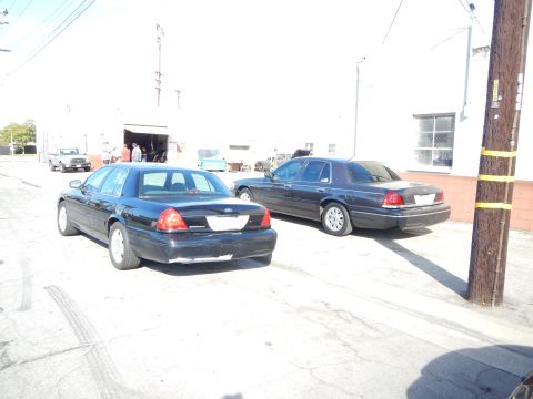 2003 Ford Crown Victoria - Two dark full-size sedans parked on a sunlit asphalt lot near an open garage door and white industrial buildings.