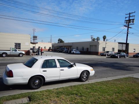 2003 Ford Crown Victoria - white full-size Ford sedan parked along a grassy curb in an industrial area with warehouses, power poles and a pickup behind.