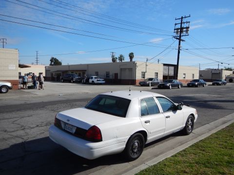 2003 Ford Crown Victoria - Rear three-quarter view of a white Ford sedan with a California plate parked beside a grass strip in an industrial lot.