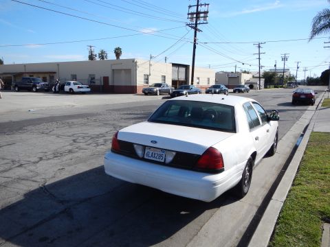 2003 Ford Crown Victoria - Rear view of a white Ford sedan with California plate 6LAX285 parked at a curb with warehouses and power poles behind.