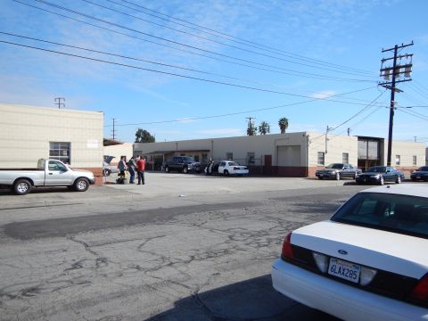 2003 Ford Crown Victoria - People gather in an industrial lot beside a white pickup and white Ford sedan with long warehouse buildings in the background.
