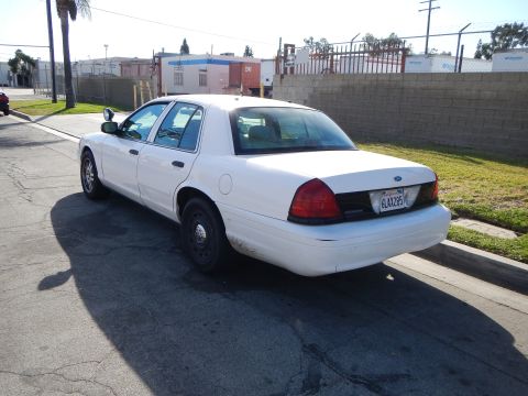 2003 Ford Crown Victoria - white Ford sedan shown rear three-quarter parked on asphalt beside a chain-link fence, curb and palm tree.