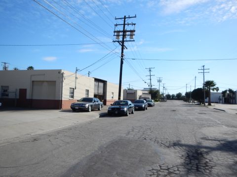 2003 Ford Crown Victoria - wide industrial street with dark sedans parked beside a beige warehouse, a tall power pole and palm trees under blue sky.