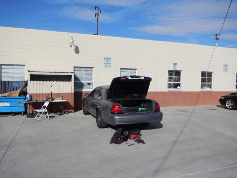 2003 Ford Crown Victoria - gray full-size sedan with its trunk open parked behind a warehouse, with a folding table, chairs and a dumpster nearby.