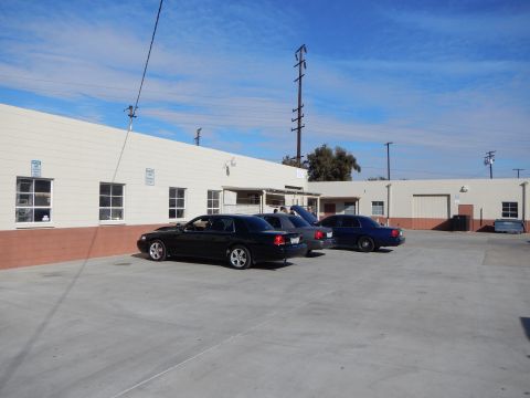 2003 Ford Crown Victoria - Three dark full-size sedans parked nose-in along a warehouse wall in a concrete lot under a bright blue sky.