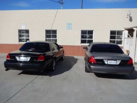 2003 Ford Crown Victoria - black and a gray Ford sedan parked nose-in against a warehouse wall, shown from the rear with California plate 6NUF092.