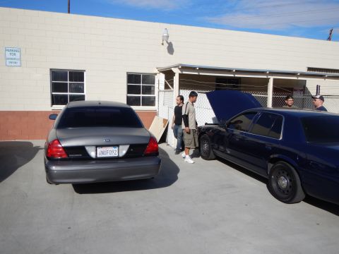 2003 Ford Crown Victoria - gray Ford Crown Victoria sedan is parked next to a dark blue car with its hood raised as men stand by an industrial building.