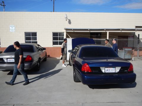 2003 Ford Crown Victoria - man in black walks between a gray and a dark blue Ford sedan parked outside a tan commercial building under a clear sky.