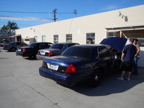 2003 Ford Crown Victoria - Several dark Ford Crown Victoria sedans line up along a tan building, the nearest blue one having its hood open with men beside it.