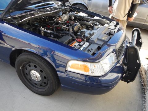 2003 Ford Crown Victoria - open front of a blue Ford Crown Victoria shows its engine bay, headlight, and black steel wheel beside a push bar.
