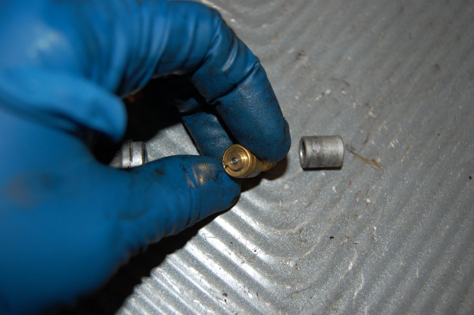 Blue gloved hand holding a brass carburetor jet next to a metal sleeve on a textured gray surface