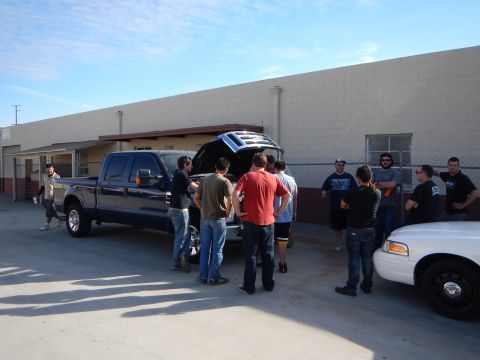 2003 Ford Crown Victoria - group of people stand around a dark crew-cab pickup truck with its hood raised in an industrial parking lot.