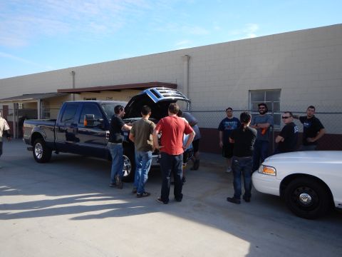 2003 Ford Crown Victoria - People cluster around a dark crew-cab pickup with its hood open next to a white sedan outside a tan building.