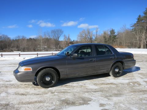 2001 Ford Crown Victoria - A dark gray 2001 Ford Crown Victoria sedan is parked on a snowy lot under a bright blue sky.