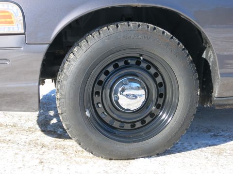 2001 Ford Crown Victoria - The front wheel and fender of a Ford Crown Victoria, showing a 'Snow Tracker' tire on a black steel rim with a Ford logo center cap.
