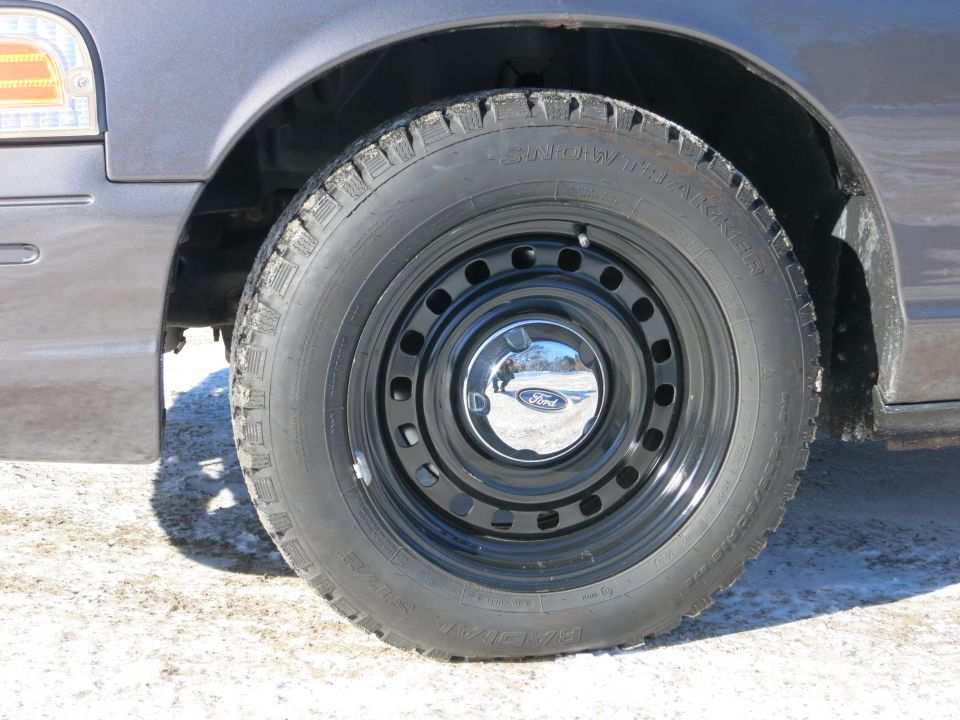Close-up of a 2001 Ford Crown Victoria's front wheel and fender. It has a black steel wheel with a chrome Ford hubcap and a 'Snow Tracker' tire