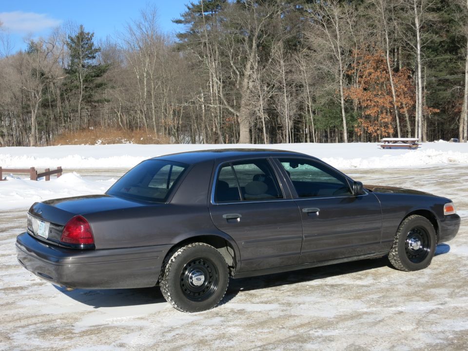 Dark grey 2001 Ford Crown Victoria sedan, rear-left quarter view, parked on snow-covered ground with trees in background