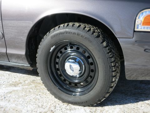 2001 Ford Crown Victoria - The front wheel of a gray 2001 Ford Crown Victoria is visible, equipped with a black steel rim and a chrome hubcap displaying the Ford logo.