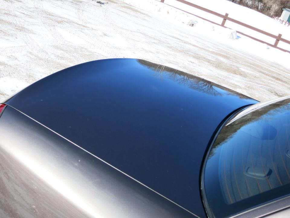 Dark car roof and rear window with snow-covered ground and fence in background