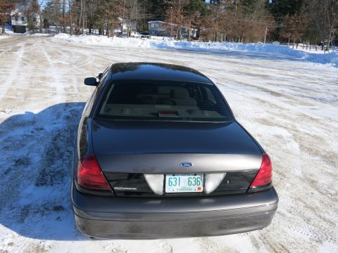 2001 Ford Crown Victoria - The rear of a dark gray 2001 Ford Crown Victoria is visible, parked on a snowy road with houses in the background.