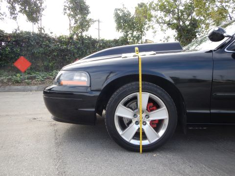 2003 Ford Crown Victoria - yellow tape measure is held vertically over the front wheel of a black sedan with a silver wheel and red brake caliper.