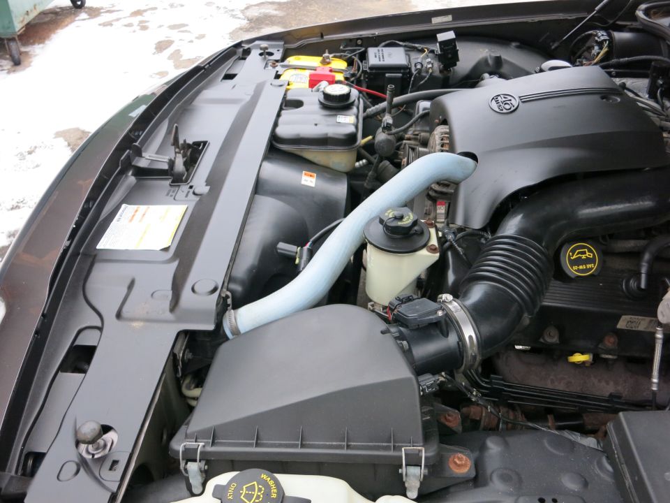 Engine bay of a car, showing the air intake, battery, and hoses. The engine cover has a circular emblem
