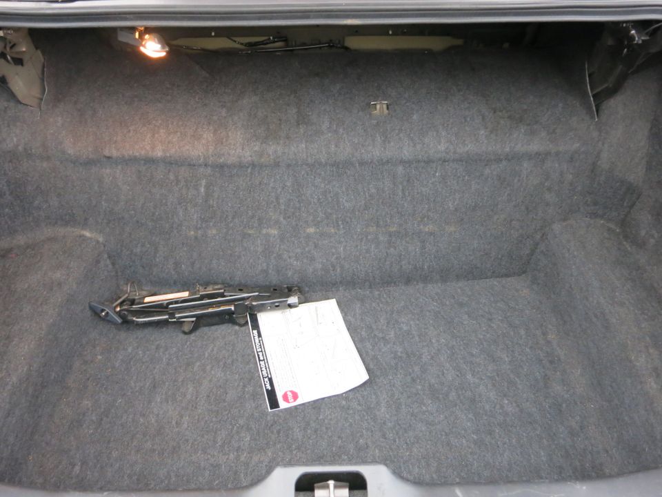 Car trunk interior with gray carpet, a scissor jack, and a folded document. A light is visible in the upper left