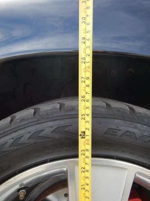 2003 Ford Crown Victoria - yellow tape measure extends vertically over a tire and dark fender, with tire sidewall lettering visible.