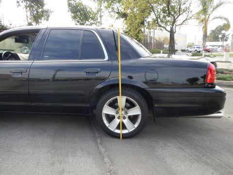 2003 Ford Crown Victoria - yellow tape measure stands vertically at the rear wheel of a black sedan parked on pavement near trees.
