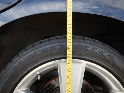 2003 Ford Crown Victoria - tape measure runs vertically from the wheel arch to a silver alloy wheel on a dark car, measuring fender-to-tire clearance.