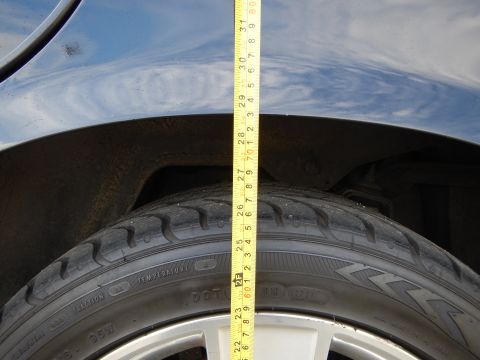 2003 Ford Crown Victoria - Tape measure spanning a dark wheel arch down to the tire and silver alloy wheel, measuring fender clearance.