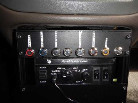 2003 Ford Crown Victoria - Hornblasters.com control panel with labeled toggle switches lit red, blue, and amber, mounted beneath a tan dashboard.