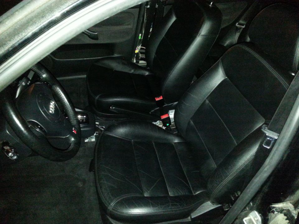 Car interior showing front black leather seats, steering wheel, and dashboard area. Some wiring is visible
